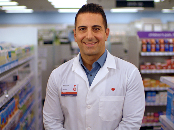 A pharmacist standing in a pharmacy aisle, wearing a lab coat.